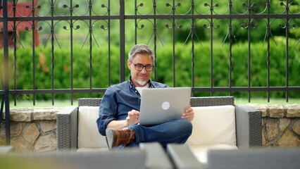 Happy middle aged man sitting on outdoor sofa in garden working with laptop computer in home office. - 516073610
