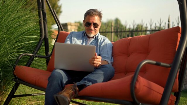 Man Working At Home Office, Sitting In Swinging Couch, Using Laptop Computer. Happy, Smiling, Relaxing. Slow Motion Video.