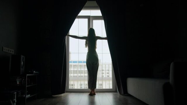 A young woman in the complete darkness of the apartment opens the blackout curtains in the morning