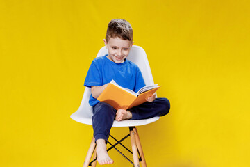 Little cheerful blond green-eyed boy 5-6 years old in a stylish blue T-shirt holding book and reading on yellow wall background, children's studio portrait