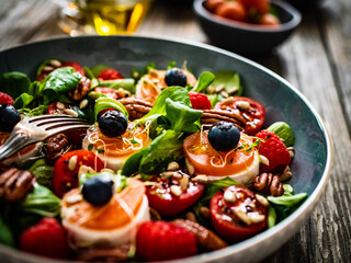 Fresh salad - goat cheese, raspberries, blueberries, pecan nuts, cherry tomatoes, pickled radishes and leafy greens on wooden table
