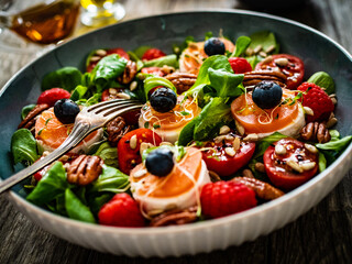 Fresh salad - goat cheese, raspberries, blueberries, pecan nuts, cherry tomatoes, pickled radishes and leafy greens on wooden table
