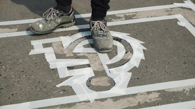 Close-up of manual utility male worker stepping on disabled car parking lot sign outlined with white tape, disability symbol, wheelchair painting on road asphalt surface, summer daytime.
