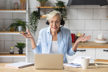 Attractive senior woman working at home in the kitchen using her laptop for conference call. Concept of mature female using internet, modern technology for learning, shopping, freelance job