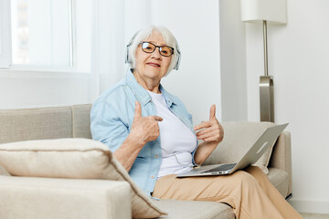 a cheerful old lady is having a video conversation with her loved ones while at a distance holding a laptop on her lap listening to them through headphones showing a thumbs up