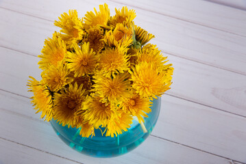 Top view on spring meadow flowers bouquet in blue water in glass vase. Dandelionson white wooden...
