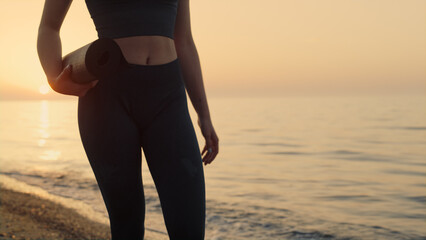 Fit girl holding mat walking seashore at sunset. Sportswoman going to training.