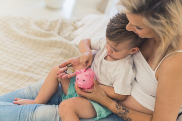 Young caucasian millennial blonde mother in a white crop top holding her little son on her laps and teaching him how to save money using piggybank. High quality photo