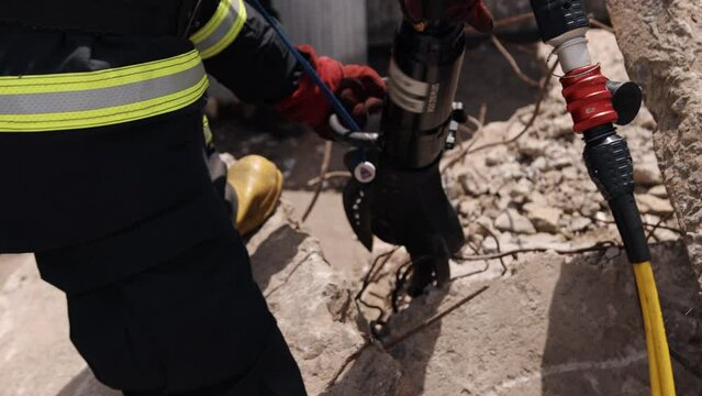 A man in a uniform rescues an earthquake victim, uses a power hydraulic cutting tool and hydraulic clamps, and is a firefighter