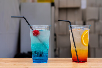 Close up of two fruity cocktails in transparent disposable glasses with straws for sale at the...