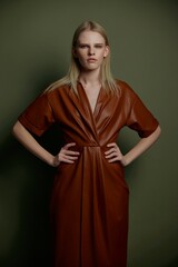 Fototapeta premium Self-confidence blonde model with blue eyes and evening makeup posing in trendy leather fashion brown palette clothes holding hands on waist looks at camera staying isolated on green background