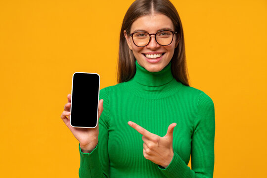 Happy young woman pointing to black phone screen, isolated on yellow background