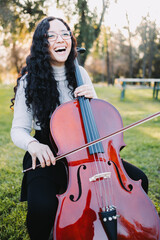 Happy young brunette woman with glasses laughing and playing cello at sunset in the park. Vertical