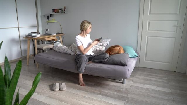 Middle-aged 45s blond woman holding smartphone taking photo of her beloved dog lying on sofa, mature female living alone spending time with domestic pet on weekend at home. Dog-human relationships 