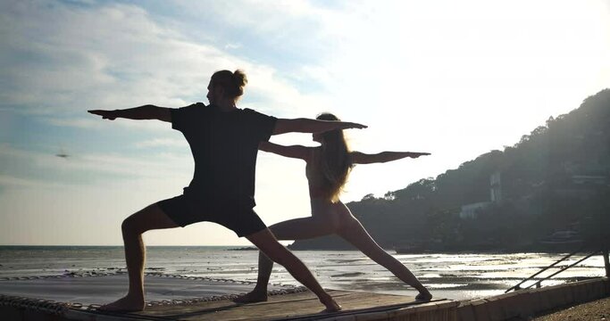 Silhouette of a caucasian man and woman with long hair finding inner peace by doing asana yoga exercises on beach at beautiful sunrise background. Healthy and Wellness lifestyle. Meditation concept.