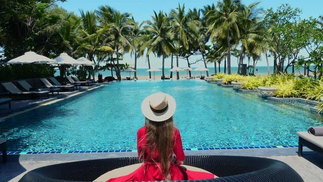 Luxury beach vacation in tropical beach hotel. Tourist woman in red dress and straw hat relax near blue swimming pool in modern resort. Female traveler on sea vacation.