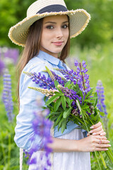 Beautiful young girl in a white dress, straw hat with a bouquet of violet flowers in her hands and picnic basket. Pretty woman in summer in the blooming field holding a bunch of purple lupin