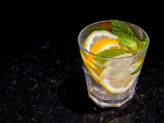 Citrus and mint leaves infused water, cocktail, lemonade in glass. Summer iced cold drink with lemon, lime, orange, mint on black marble background, copy space, side view