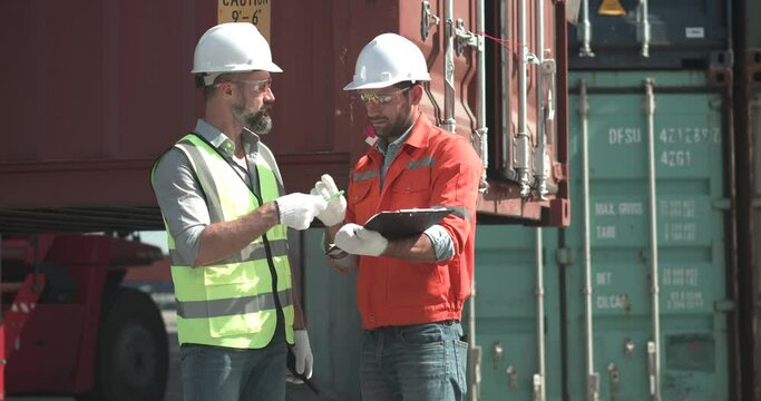 Foreman in hardhat, safety vest and glasses sign an import export document for male engineer. They shaking hands with a smile at warehouse logistic in cargo freight. Shipping and transport concept.