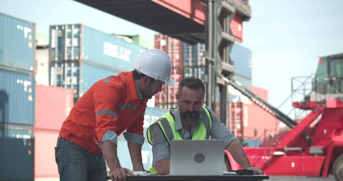 Male engineer and Foreman in safety vest using laptop computer to check control loading Containers box and use radio/walkie talkie to contact the other on site at warehouse logistic in cargo freight.