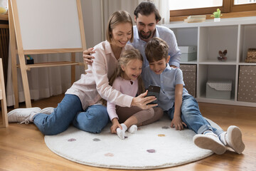 Happy excited parents and little sibling kids talking on video call, using smartphone together, talking selfie photo, laughing, feeling joy, having fun, sitting on heating floor at home