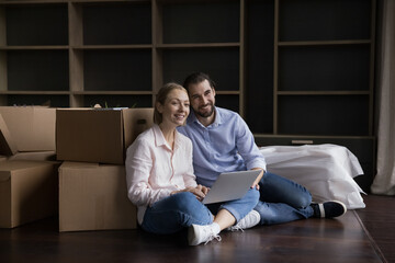 Happy couple using laptop, consulting internet for interior design samples, sitting at stacked cardboard boxes on floor, using laptop, ordering shipping service, smiling, laughing, looking away