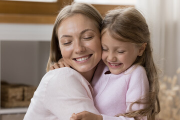 Happy loving mom hugging sweet little preschool daughter girl with closed eyes, cheek touches, toothy smile. Young mother cuddling kid with care, tenderness, devotion, enjoying motherhood