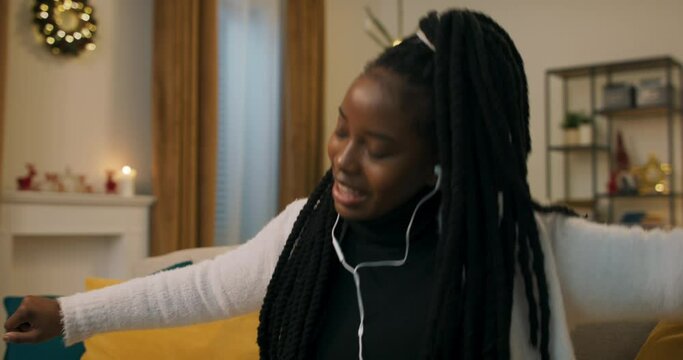 Close-up of attractive girl of African appearance who listens to music on headphones. The girl is in good mood: she sings, waves arms, moves in time and smiles. A Christmas tree flickers in the room.
