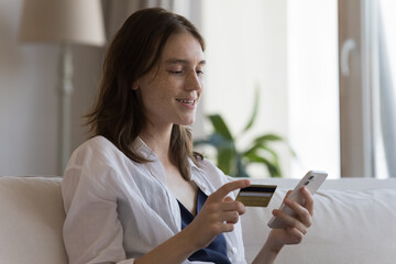 Freckled young woman sit on sofa at home holds credit card and cellphone buy goods via web store...