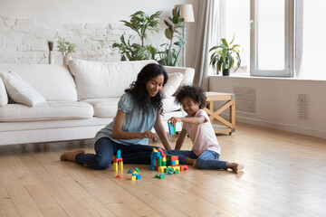 African mother and little girl play wooden colorful cubes, build towers sit on warm floor in living...