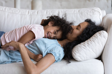 African adorable little daughter cuddles her mother lying resting on cozy couch at home, spend...