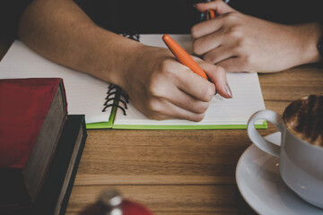 Woman hand with orange pen writing on notebook at coffee shop. Woman working in coffee shop.