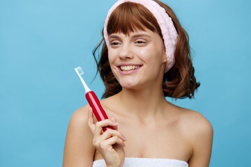 a joyful, pretty woman holds an electric red toothbrush in her hand, standing on a blue background and looking at the camera, smiling broadly with clean, light teeth, holding her hand near her face