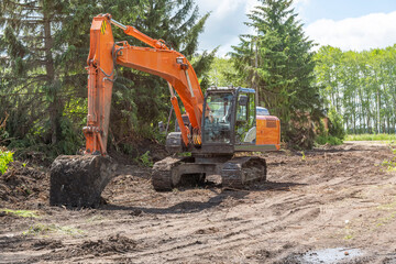 Large orange crawler excavator stands idle against backdrop of green fir trees and blue cloudy summer sky.