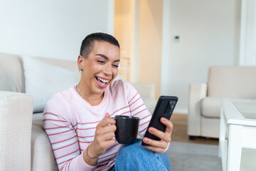 A young woman in a sweater with a cup in her hands looks at the phone while sitting on the couch.