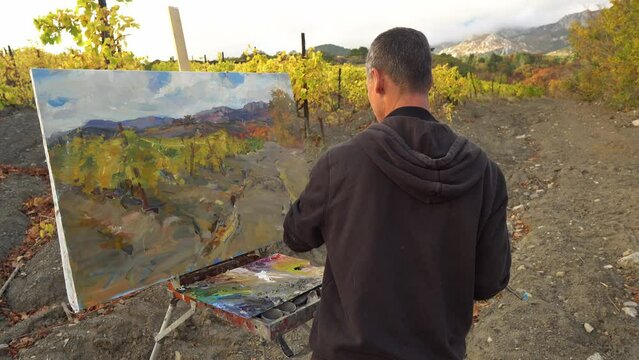 Man artist in front of easel outdoors, painting with brushes, using oil or acrylic
