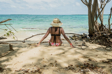 Mujer con sombrero y bikini sentada en la arena mirando el horizonte en la playa de una isla en el...