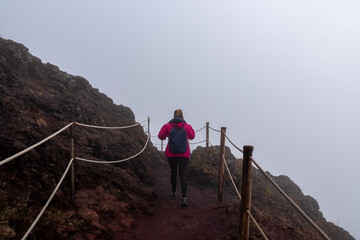 Tourist woman hiking along foggy edge of active volcano crater of Mount Vesuvius, Province of Naples, Campania region, Italy, Europe, EU. Volcanic landscape full of stones, ashes and solidified lava