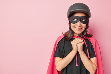 Horizontal shot pleased woman keeps hans together looks with dreamy expression aside wears helmet mask black t shirt and cape pretends being superhero imagines something poses indoors pink wall