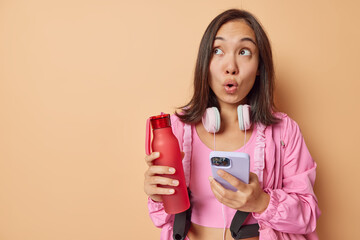 Surprised Asian woman rests after workout holds mobile phone and bottle with water looks amazed...