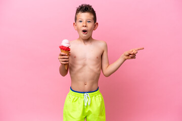 Little caucasian boy eating an ice-cream isolated on pink background surprised and pointing side