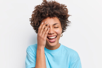Fototapeta na wymiar Horizontal shot of cheerful woman with curly hair makes face palm smiles broadly expresses positive emotions dressed in casual blue t shirt isolated over white background hears something funny