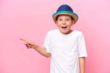 Little caucasian boy wearing a hat isolated on pink background surprised and pointing finger to the side