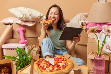 Cheerful Asian woman enjoys eating pizza and watching film on portable tablet poses on floor around...
