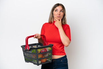 Young Rumanian woman holding a shopping basket full of food isolated on white background having doubts