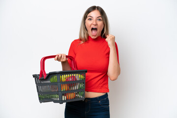 Young Rumanian woman holding a shopping basket full of food isolated on white background celebrating a victory in winner position