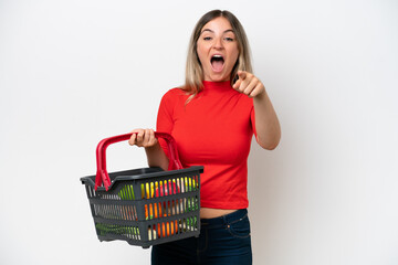 Young Rumanian woman holding a shopping basket full of food isolated on white background surprised and pointing front