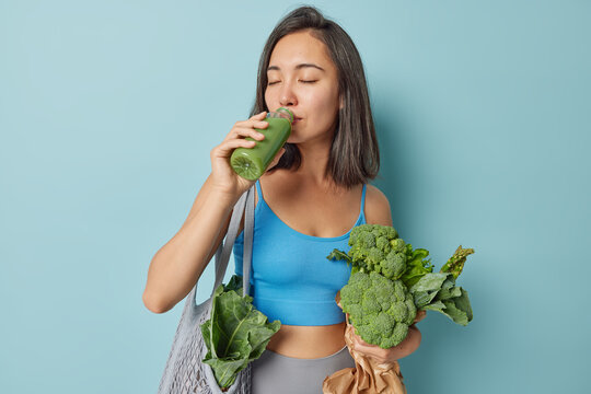 Dark haired slim Asian woman keeps to healthy diet drinks homemade fresh green smoothie carries vegetables in net bag isolated over blue background. Vegan meal super foods and detox concept.