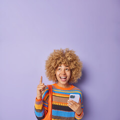 Pretty positive young woman has natural curly hair points index finger above laughs with excitement demonstrates awesome promotion wears knitted striped jumper isolated over purple background