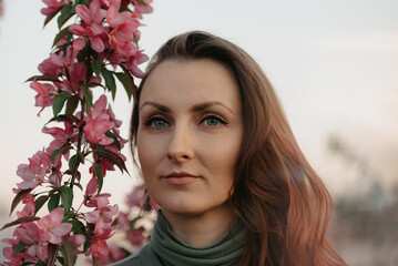 A headshot of a redhead woman is staring into the distance near flowering sakura at sunset.